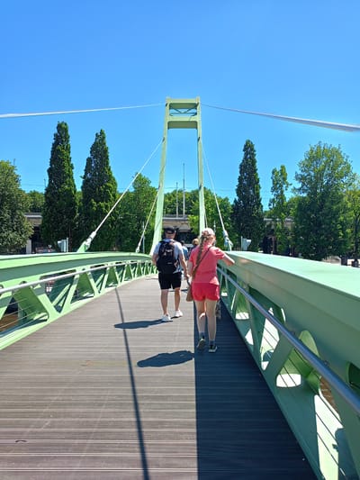 The image depicts a scene on a modern pedestrian bridge with a green metal structure, likely in Kaliningrad, Russia. Two people are walking away from the camera across the wooden walkway. The person on the left is a man wearing a backpack, black shorts, and white sneakers. The person on the right is a woman wearing a pink t-shirt, red shorts, and white sneakers, carrying a small bag over her shoulder. The bridge has a cable-stayed design, with a prominent green pylon in the background supporting cables. Tall, slender trees line the sides of the bridge and extend into the background, suggesting a park-like or waterfront setting. The sky is clear and blue, indicating a sunny day, likely midday due to the strong shadows cast on the bridge. The overall mood appears casual and pleasant, with people enjoying an outdoor stroll. No visible text is present in the image.