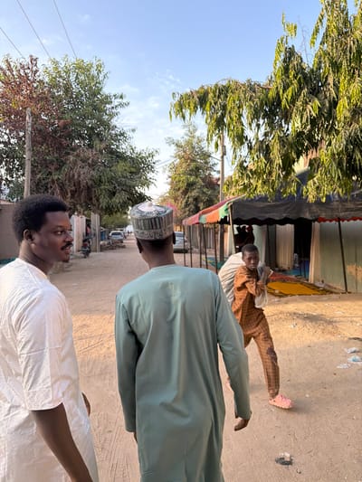 The image depicts an outdoor street scene in Daurawa, Nigeria, under a clear, bright sky. Three male individuals are prominent in the foreground and midground, walking on an unpaved, light brown dirt road.
In the left foreground, a dark-skinned male with short, curly dark hair and a mustache is visible from the chest up, oriented in profile, looking towards the right side of the frame. He wears a light-colored, long-sleeved garment, possibly white or cream-colored, with a textured pattern.
Adjacent to him, positioned slightly further back and towards the center, another dark-skinned male is seen from behind, wearing a long-sleeved, light green tunic and a white, green, and dark patterned cap. He is walking away from the camera, down the street.
In the midground, to the right of the person in green, a younger dark-skinned male is walking towards the right, carrying a large, light-colored sack or bundle over his left shoulder. He wears a brown and tan checkered long-sleeved shirt and matching trousers, along with pink Crocs-style footwear. He is looking directly towards the camera.
The environment includes several large trees with green foliage, particularly a prominent one on the right with elongated leaves and another on the left with reddish-brown leaves. Utility poles and overhead wires are visible against the blue sky. On the right side of the street, a temporary market stall or structure is present, featuring a dark tarp roof, a colorful striped valance (red, green, orange), and light-colored fabric walls. Inside this structure, yellow and other colored rugs or textiles are visible. Further down the street, in the background, other buildings and parked vehicles, including what appears to be a motorcycle, are discernible. The ground is dusty and appears dry, with some scattered debris.