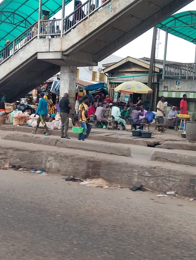 This media file depicts a bustling street scene in Shangisha, Nigeria, likely during daylight hours given the illumination. The foreground is dominated by a road with a raised concrete curb, on which several individuals are present. A man in a blue shirt and patterned shorts walks from left to right, his gaze directed forward. To his right, a man in a black shirt and grey pants stands facing away from the camera. Another man, wearing a yellow and black striped shirt, walks from right to left, carrying a green basket.
In the midground, a covered pedestrian walkway stretches across the frame. People can be seen walking on the walkway, some carrying items. Below the walkway is a market area. Stalls are set up, some with goods displayed, and several people are gathered, possibly shopping or socializing. A yellow umbrella provides shade over one of the stalls. Various objects are visible, including baskets of produce, bags, and what appear to be food items on a table. The environment suggests an active commercial hub.
The background reveals more of the market area and buildings. The overall atmosphere appears busy and typical of a Nigerian marketplace, with a mix of shoppers, vendors, and pedestrians navigating the space. The weather appears to be fair, with no visible signs of rain. The scene is vibrant with human activity, highlighting the everyday life in this location. There is no visible text in the provided image.