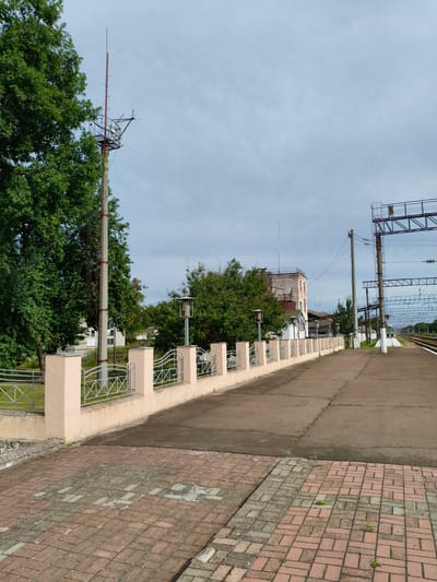 The image depicts a view of a train station platform in Hoholeve, Ukraine. The platform is made of concrete and is lined with a decorative fence with beige pillars. To the left, there is a large green tree and a tall pole with a radio antenna. In the background, there are several buildings, including a brick building with a white facade. Train tracks are visible in the distance on the right side of the image, with overhead electrical lines. The sky is overcast. The scene is still and appears to be devoid of people.