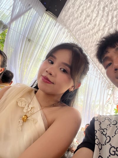 This image captures a close-up, upward-angled perspective of two individuals and their surroundings, likely a selfie taken from below. The primary subject is a young woman with dark hair, looking directly at the camera with a gentle smile. She is wearing a light-colored, possibly cream or pale yellow, strapless or off-the-shoulder dress, featuring delicate floral or ruffled appliques on the bodice. A fine gold chain necklace with a prominent, round gold pendant rests on her collarbone.

Above and slightly behind her, the upper part of a man's face – including his forehead, dark hair, and eyes – is visible. He is wearing a white shirt with a repeating black "G G" pattern, resembling a well-known designer logo.

The setting appears to be an outdoor or semi-outdoor event, with a canopy of draped white sheer fabric creating a ceiling effect. Bright, diffused light filters through these drapes, indicating daytime. Green foliage and hints of natural light are visible beyond the translucent fabric, suggesting a garden or park venue. In the lower background, other blurred individuals and their heads can be seen, further suggesting a social gathering. A dark panel with small white lights is visible in the upper left corner, possibly part of a screen or decoration.

The overall atmosphere suggests a festive or celebratory event in Bình An, Vietnam, possibly a wedding or party, given the attire and decorative canopy. The direct gaze of the woman conveys engagement and a sense of personal connection to the camera.
