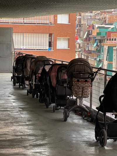 In the city of Santa Coloma de Gramenet, Spain, a row of strollers is parked on a concrete surface. The strollers are lined up along a metal railing with a wire mesh. Behind the railing, multi-story buildings with balconies and awnings are visible, suggesting an urban environment. The ground is wet and reflective, with shadows cast by the strollers and the overhead structure. The scene appears to be an outdoor parking or storage area for baby strollers, possibly in a residential or community setting. No people are visible in the image.