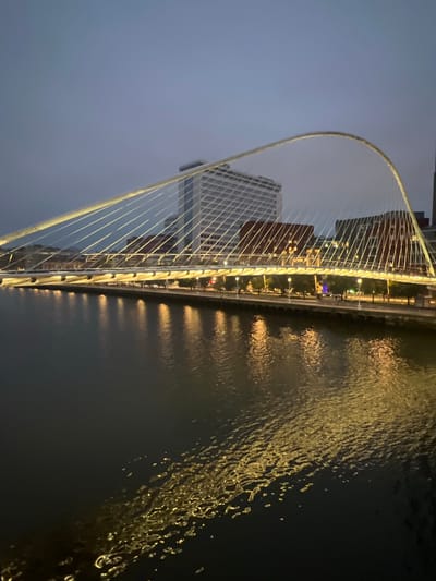 This is a photograph of the Zubizuri bridge in Bilbao, Spain, taken at dusk. The cable-stayed bridge, designed by Santiago Calatrava, spans the Nervión River. The white pedestrian bridge features a distinctive curved mast and a deck supported by white cables. The bridge is illuminated with warm yellow lights, which cast reflections on the dark water of the river below. In the background, several modern buildings are visible, including a tall, white skyscraper with a grid-like facade and a red brick building. Trees line the riverbank in front of the buildings. The sky is a deep blue, transitioning to a lighter hue towards the horizon, indicating the twilight hour. There are no people clearly visible in the image, and no specific actions or interactions are depicted. The overall scene conveys a sense of urban tranquility and architectural beauty in the evening light.