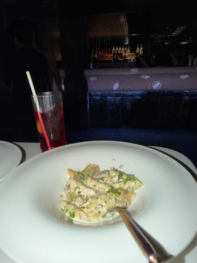 This image depicts a dimly lit indoor dining and bar area in New Delhi, India. In the foreground, a white, wide-rimmed ceramic plate contains a serving of penne pasta in a light-colored, creamy sauce, garnished with chopped green herbs. A dark metallic utensil rests within the pasta. To the left of the pasta plate, a portion of a second white plate is visible. A tall, clear glass, exhibiting condensation, holds a red beverage with a white plastic straw and a visible slice of yellow citrus fruit.
In the midground, to the left, the dark silhouette of an individual, possibly a diner, is partially visible. The background features a bar counter with a dark, textured base. The top surface of the bar counter is illuminated, displaying projected abstract symbols, including eye-like and oval shapes. Behind the bar, numerous liquor bottles are arranged on shelving, catching ambient light from the dark environment. Indistinct dark figures, consistent with staff or patrons, are present behind the bar and further in the background.