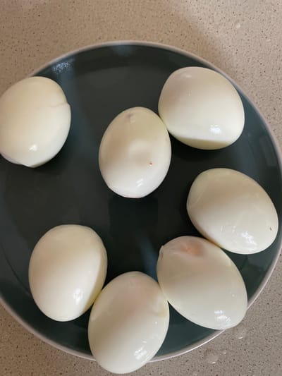 Seven peeled hard-boiled eggs are arranged on a dark grey plate. The plate is placed on a speckled countertop. The eggs are white and have a smooth, slightly wet appearance, suggesting they have recently been peeled. One egg on the right side of the plate shows a slight pinkish hue, possibly from being cooked with the shell or some residue. The lighting appears to be natural, casting subtle highlights on the eggs. The scene is a close-up shot, focusing entirely on the eggs and the plate. There are no people visible, and the activity taking place is simply the presentation of the prepared eggs on a plate. The setting is a kitchen or food preparation area, indicated by the countertop. There is no visible text in the image.