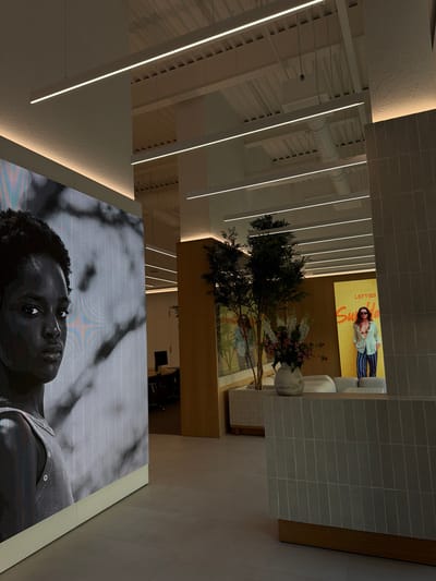 The image depicts the interior of a modern commercial space, likely a store or office lobby, with a minimalist aesthetic. Prominently featured on the left is a large, high-resolution digital display showcasing a black and white portrait of a young person. The display is illuminated and casts a soft light on the surrounding area. To the right, there is a section of tiled wall, likely a reception desk or architectural feature, with a wooden base. Further to the right, a bright yellow advertisement poster is visible, featuring a person wearing sunglasses and stylish clothing, with text that appears to read "LEFTIES" and "Summer". In the middle ground, there is a potted artificial tree with dark green foliage, and a vase of flowers. Behind the tree, a beige wall with subtle patterned wallpaper is visible. Beyond that, there are glimpses of office furniture, including chairs and desks, suggesting a functional workspace. The ceiling is equipped with multiple linear LED light fixtures, providing ambient illumination. The floor appears to be tiled with large, neutral-colored tiles. The overall impression is one of a clean, contemporary, and inviting environment. There is no identifiable signage or context to suggest the location is in Fibracolor, Spain.