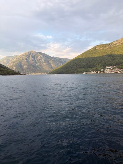 The image depicts a wide expanse of dark, rippling water in the foreground, likely a bay or fjord. In the background, verdant, tree-covered hills rise steeply on either side, meeting a hazy, overcast sky. Nestled on the slope of the right-hand hill is a cluster of white-roofed buildings, suggesting a coastal village. The architecture of the buildings, with their red-tiled roofs and white walls, is characteristic of Mediterranean settlements. A small sailboat is visible on the water near the shore. The lighting suggests either early morning or late afternoon, with shadows cast by the hills and a soft glow on the peaks. The overall impression is one of natural beauty and tranquility. While the location is specified as Đurići, Montenegro, the image itself does not contain any identifying landmarks or signs to confirm this specific location; it is a generic depiction of a coastal mountainous landscape.