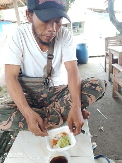 A man in a black baseball cap, white t-shirt, and camouflage pants sits cross-legged, eating from a white container. He uses a plastic spoon in his left hand and a pen in his right. A small cup of dark liquid is nearby. He wears a strap across his chest. Behind him are a blue barrel and wooden benches. The timestamp "Vivo Y19s Akthar & Arvan 10 Des 2025, 14:15" is visible in the lower left corner.