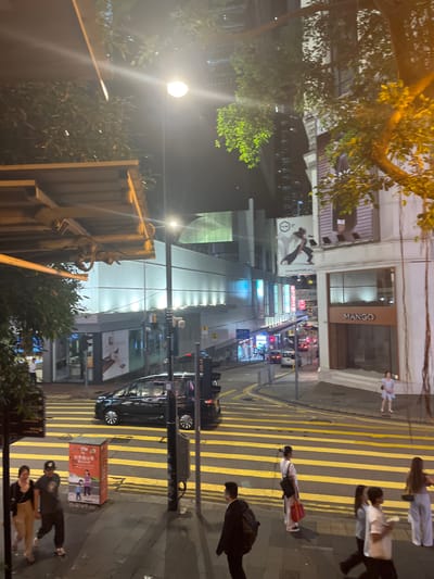 A nighttime urban scene in Hong Kong, China. Pedestrians are crossing a street marked with yellow stripes. A Mango store is visible in the background along with billboards. Cars are driving on the street. A street light illuminates the scene.