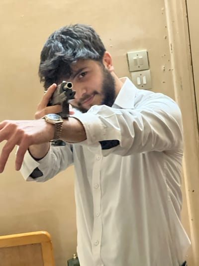 A man with dark hair and a beard is holding a gun, pointed at the camera. He is wearing a white dress shirt with a gold watch on his left wrist. In the background, there is a beige wall with two light switches and a portion of a wooden desk with rounded edges. The photograph is taken from a low angle, looking upwards at the man, and appears rotated.