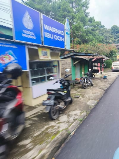 The image shows a street scene in Lembang, Indonesia, located at coordinates -6.823 latitude and 107.61 longitude. The street is paved and runs diagonally from the left foreground to the right background of the image. On the left side of the street are several buildings, one of which features a large sign advertising a product called "Warnas Ibu Ocih." There is a small shop with a glass front and a storefront sign, "100% MURNI." Two motorcycles are parked on the side of the street, and another motorcycle is partially obscured by a building. The right side of the street is lined with a row of low-lying, dilapidated buildings with open sides. In the background, a white van is visible, parked on the right side of the road. There is a green tree line in the background, and electrical wires run diagonally across the image from the left foreground to the right background. There are no people visible in the image.