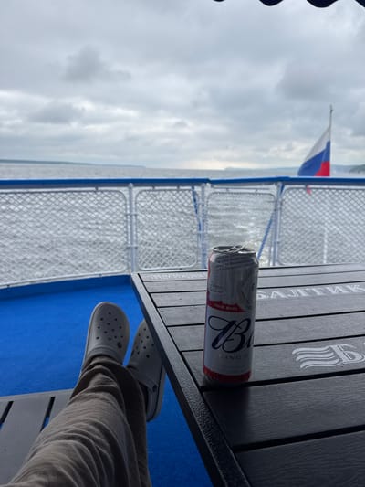 An individual, wearing light-colored Crocs and beige pants, is seen from the knees down, relaxing on a boat. Their feet are propped up on a dark-colored lounge chair. A can of Budweiser beer rests on a dark wooden table in front of them. The table has a logo with the letters "Б" and wavy lines etched into it, and text that appears to be in Cyrillic. In the background, a body of water stretches to the horizon under a cloudy sky. A Russian flag is visible on a flagpole in the distance. The boat's railing, with a white woven pattern, separates the deck from the water. The deck itself is a vibrant blue. The scene is set in the city of Yashel'cha, Russia.