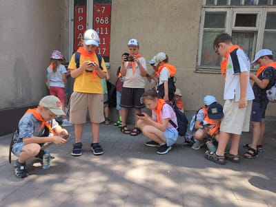 A group of children, mostly wearing orange scarves, are gathered outside a building in Novorossiysk, Russia. The building appears to be old, with a textured wall and a boarded-up window. Some of the children are looking at their phones, while others are standing or squatting. One child in the foreground is wearing a blue and white baseball cap and a yellow t-shirt. Another child next to him is wearing a grey t-shirt and a tan baseball cap. A bottle of water is on the ground next to the child in the grey t-shirt. The overall scene suggests a casual gathering, possibly a break during an outdoor activity or excursion. The presence of the building and the paved ground indicates an urban or semi-urban setting.