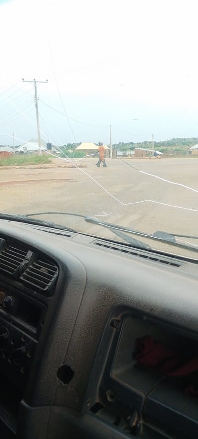 This image captures a scene from inside a vehicle in what appears to be the city of DU, Nigeria. The foreground is dominated by the interior of the vehicle, showing a portion of the dashboard with air vents and a dark textured material. Through the windshield, a man in an orange outfit is seen walking across a dusty, unpaved road. The road is marked with white tape in several places, suggesting some form of activity or construction is taking place. In the background, there are several small structures, some with corrugated metal roofs, and a utility pole with multiple wires. The sky is overcast, and the overall impression is of a rural or developing area. The image is taken from a low angle, as if from the driver's perspective.