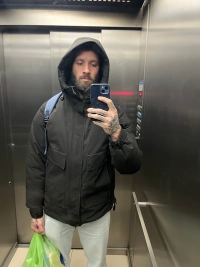 The image captures a man taking a mirror selfie inside an elevator. He is seen from the chest up, oriented sideways in the frame, and is wearing a dark hooded jacket and light-colored pants. A blue strap from a shoulder bag is visible beneath his jacket on his left side. The man has a beard, and prominent tattoos cover his hands. He holds a dark-colored smartphone in his right hand, reflecting his face and the elevator interior. In his left hand, he holds a bright green plastic bag.
The setting is an elevator car, characterized by polished metallic (likely stainless steel) walls and vertical paneling. On the right side of the elevator, a metallic handrail is visible. On the left, a darker panel with vertical slots can be seen. Above the man's right shoulder, an elevator control panel is visible with illuminated elements, including a blue symbol resembling an 'H' and several red lights, possibly indicating floor numbers or status. The man's expression is neutral. The overall impression is an indoor, everyday scene. No discernible text is clearly readable on the elevator panel or elsewhere.