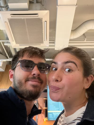 Two individuals, a male and a female, are depicted in a close-up indoor photograph, likely a selfie. The male, positioned on the left, wears dark sunglasses and a dark blue collared, zippered jacket; he has dark hair and a beard, exhibiting a slight smile. The female, on the right, has dark hair pulled back and makes a playful expression with puckered lips and wide eyes; she wears a striped shirt under a dark jacket. The background reveals an industrial-style ceiling with exposed white utility pipes and a white rectangular cassette air conditioning unit. To the right of the female, a digital menu board with light text on a dark background is partially visible, displaying Czech language, including the phrase "Přesně podle V". A light-colored slatted wall and an orange ambient light source are also present. The scene is within a commercial establishment in the Capital City of Prague, Czechia.