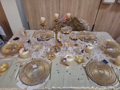 This image depicts an indoor, static display of a festive table setting, likely a retail presentation in Blainville-sur-Orne, France. A rectangular table is draped with a cream-colored tablecloth featuring a gold glitter effect and subtle white snowflake or star patterns. The table is arranged for multiple place settings, each comprising a large, clear glass charger plate with a gold textured pattern, topped by a smaller, matching plate. Gold-colored forks are positioned to the left of the plates, and gold-colored knives to the right.
Central to the table is a three-armed golden candelabra holding three lit, cream-colored pillar candles. Various decorative elements, primarily in gold and clear glass, are distributed across the table. These include multiple glittering golden apple-shaped ornaments, some with attached white retail tags, and small golden Christmas tree figurines. Several clear stemmed wine glasses and champagne flutes are present. Other objects include clear glass bowls or decorative vessels, some with gold rims, and scattered small golden star confetti on the tablecloth. In the upper left quadrant, a bottle, possibly of spirits, wrapped in clear plastic, stands near a clear glass serving dish. The background consists of light brown wooden wall paneling. No individuals are visible in the scene.