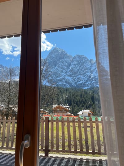 The image shows a view from a window in Sappada, Italy. Outside the window is a snow-covered mountain, a dense evergreen forest, wooden buildings with orange and white accents, and a grassy area enclosed by a wooden fence. A curtain hangs in the upper left of the frame. The window itself is made of brown wood and has a metal handle.