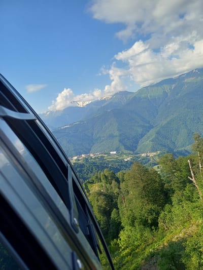 The image captures a scenic mountain landscape from the perspective of looking out of a vehicle, possibly a car or cable car. The foreground is dominated by the dark, metallic frame and roof structure of the vehicle, with visible hinges and latches. The scene extends into a wide vista of lush green mountainsides densely covered in trees. In the distance, snow-capped peaks rise above the treeline, hinting at a higher elevation. A small town or village with white buildings is nestled within the valley, appearing almost miniature against the grand scale of the mountains. Patches of bright blue sky with scattered white clouds are visible, suggesting fair weather and daytime. The sunlight casts a warm glow on the scene, highlighting the textures of the foliage and the contours of the mountains. The orientation of the image is rotated, as if the camera was tilted to capture the expansive view. The overall impression is one of natural beauty and a sense of journey or observation from a moving viewpoint.