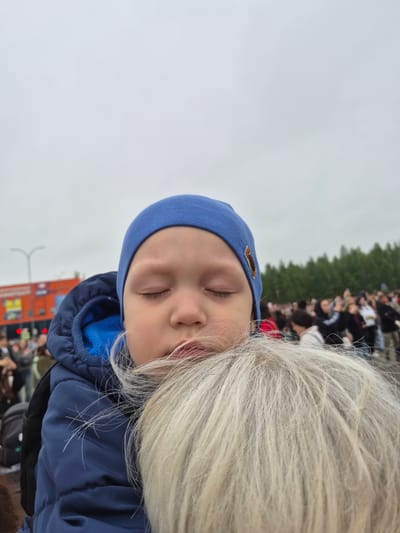 The image captures a close-up of a young child with eyes closed, wearing a blue beanie and a blue jacket, being held by an unseen person. The child's face is partially obscured by strands of the holder's light-colored hair. In the background, a crowd of people is visible, with trees and a building with an orange facade featuring red lights in the distance. The setting appears to be an outdoor event in Nyagan, Russia, suggested by the mention of the city. The overall atmosphere is somewhat overcast.