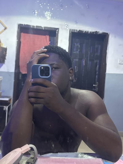 A dark-skinned individual with curly hair is taking a selfie in what appears to be a bathroom. The individual is holding a blue iPhone in front of their face, obscuring part of it. They have an earring in their left ear. The background includes a doorway with a dark interior and a red object visible inside. A mirror reflects the scene, and there are some light fixtures and other objects visible in the reflection. The location is given as Gbongan, Nigeria.