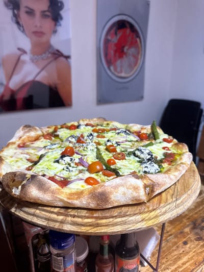 A pizza with tomato, onion, asparagus, and spinach toppings is presented on a wooden serving platter at a restaurant or food establishment in Chipilo de Francisco Javier Mina (Chipilo), Mexico. The pizza's crust is thick and slightly charred. The serving platter sits on a wooden surface beside various bottles and containers. A framed picture of Elizabeth Taylor and a framed picture of a round, reddish object (possibly a sliced tomato) are displayed on the wall.