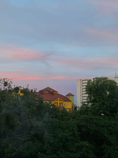 The image depicts a city skyline at dusk, with a gradient of pastel colors in the sky. In the foreground, lush green trees dominate the lower half of the frame. Behind the trees, a bright yellow building with a red roof is visible, featuring multiple windows and architectural details. To the right of this building, a tall, modern white high-rise with grey accents stands out. The overall scene suggests a residential or mixed-use area. The provided information indicates that the location is Poznan, Poland, however, without specific landmarks or context, pinpointing an exact neighborhood within Poznan is not possible based solely on the visual information.