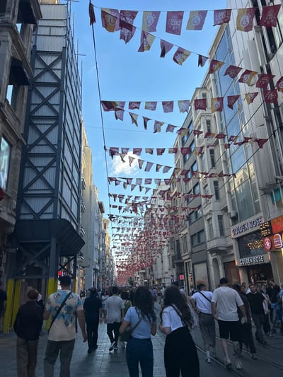 The image shows a crowded pedestrian street in Beyoğlu, Turkey, likely Istiklal Avenue. A long string of decorative flags, predominantly red and yellow with a crest and the number "26" on them, are strung across the street, creating a festive atmosphere. Buildings line both sides of the street, with some appearing to be under construction or renovation, indicated by scaffolding and covered facades. Several people are walking in both directions on the cobblestone street, which also has tram tracks embedded in it. Stores and businesses, including "GOLDEN ROSE" and "BURGER KING," are visible with their signage. The sky is blue with some clouds, and the overall lighting suggests it is daytime. The people are dressed in casual summer attire.