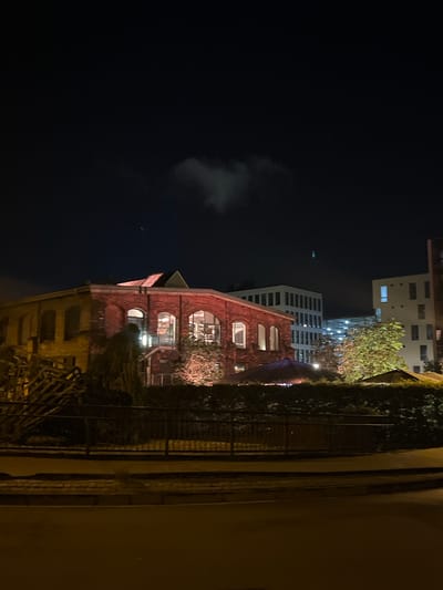 The image captures an urban night scene, predominantly dark with artificial lighting illuminating various structures.
**Subjects:**
The central subject is a multi-story brick building, possibly an older industrial or renovated structure, featuring several arched windows that emit a warm, golden light from within. Trees with illuminated foliage are clustered around this building, suggesting landscaping. Behind the brick building, a taller, modern building with a light-colored facade and numerous rectangular windows is visible, also showing interior lights. Further in the background, another modern building with a lighter exterior can be seen. On the far right, a dark wall or structure, partially covered in dark ivy or climbing plants, stands next to a dark metal railing or fence.
**Activity or event taking place:**
No direct human activity or specific event is discernible. The illuminated windows suggest that the buildings are occupied or in use during the night.
**Notable details:**
The time of day is clearly nighttime, characterized by the dark sky and reliance on artificial lights. The sky, occupying the upper left portion of the image, is dark with faint, wispy clouds visible. There are no distinct weather conditions apparent beyond the clouds. The blend of classic brick architecture with modern high-rise buildings indicates an urban setting. The perspective appears to be slightly elevated, looking across towards the buildings. No visible text is present in the image. The overall mood is quiet and still, typical of a city at night.