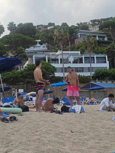 The image shows a group of people relaxing on a sandy beach in Blanes, Spain. The setting appears to be a resort area, with a multi-story building with balconies and windows visible in the background, surrounded by lush greenery and palm trees. Beach umbrellas and lounge chairs are scattered across the sand, indicating a leisure activity.
Several individuals are the main subjects. In the foreground, a young man in pink and red ombre swim trunks is standing and looking towards the camera, holding a phone. To his right, another young man, also shirtless, is sitting on a lounge chair wearing patterned swim trunks. Further to the right, a man in a white shirt and tan pants is sitting, appearing to be looking down. Other people are visible in the background, some lounging on chairs, suggesting a relaxed atmosphere.
The lighting suggests it might be late afternoon or early evening, with the sky appearing overcast. The overall mood seems to be one of vacation and relaxation. No specific text is clearly legible in the image. The presence of beach amenities and the style of architecture point to a warm, sunny climate typical of coastal Spain.