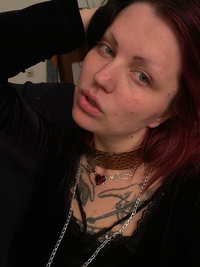 A close-up, angled shot depicts a fair-skinned individual with light green eyes and reddish-brown hair. Their hair is predominantly black on one side and reddish-brown on the other. The person is wearing black clothing with lace trim at the neckline. A prominent tattoo is visible on the upper chest and neck. Two necklaces adorn the individual: one is a wide, textured gold collar, and the other is a long silver chain necklace with a pendant of a hand holding a red gem. The background includes a glimpse of a wooden headboard and a light-colored wall with an electrical outlet. No evidence indicates the location is in Kunje, Montenegro.