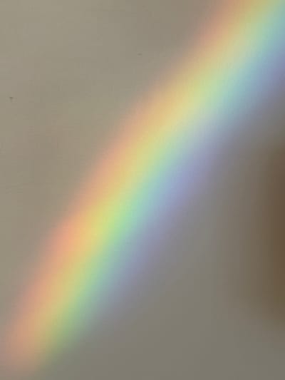 The image shows a diagonal band of light, resembling a rainbow, cast on a textured, neutral-colored surface, likely a wall. The light spectrum includes shades of red, orange, yellow, green, blue, and violet, transitioning smoothly from one color to the next. The surface itself appears to be a light beige or gray, with subtle variations in tone. There are a few small, dark specks visible on the surface, possibly dust or imperfections. The right side of the image shows a darker, blurred area, suggesting a shadow or an object out of focus. The overall scene is abstract and minimalistic, focusing on the interplay of light and color. There is no indication of the city of Vinnytsia, Ukraine, or any other specific location. The image depicts an indoor setting or an area where light is being refracted or reflected to create the colored band.
