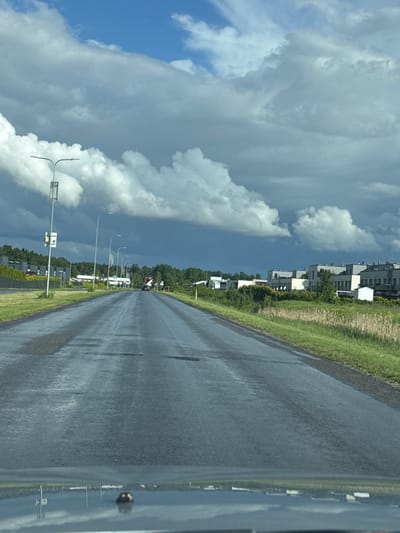 The image depicts a wet asphalt road in Tiskre, Estonia, bordered by green grass and dry vegetation. On the left side of the road, there are tall streetlights and some residential buildings with dark facades behind a fence. On the right side, a row of modern, light-colored residential buildings with multiple stories is visible, set against a backdrop of green trees. The sky is dramatic, filled with dark, heavy clouds, suggesting an impending storm, with patches of bright blue sky peeking through. The road itself is wet and reflects the sky and surrounding environment, indicating recent or ongoing rain. A dark-colored car is visible in the distance, driving down the road. The foreground shows the reflection of the sky and buildings on the surface of a car's windshield or hood.