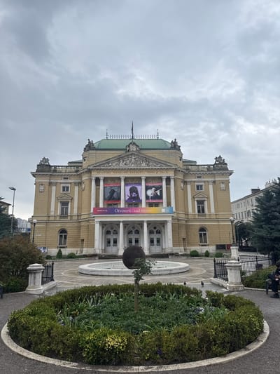 The image shows the Croatian National Theater Ivan pl. Zajc in Rijeka, Croatia. The building is a cream-colored classical structure with a green dome and ornate details. Banners advertising performances, including "Turandot", "Mali Princ", and "Balkanski Spijun" hang from the facade. A fountain and manicured gardens are in front of the building. The sky is overcast.