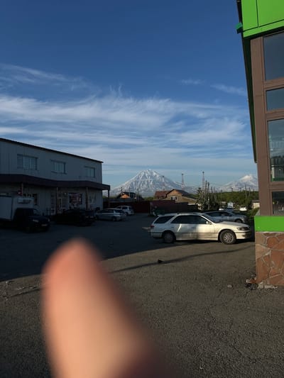 The image depicts a street scene in Yelizovo, Russia, with a prominent view of the Vilyuchinsky volcano in the background. In the foreground, a gravel parking lot is occupied by several vehicles, including a white station wagon, a black SUV, and a white truck. A partially blurred finger is visible in the lower left corner of the frame, suggesting the photo was taken by someone holding a camera or phone.

To the left, a long, single-story building with a dark roof and white walls houses a commercial establishment, possibly a shop, indicated by signage visible through its windows. A white box truck is parked in front of this building. To the right, a modern building with green and brown siding is partially visible, featuring large windows and a stone-like base.

The sky is a vibrant blue, streaked with wispy white clouds. The Vilyuchinsky volcano, snow-capped and majestic, dominates the middle ground, with other volcanic peaks visible in the distance. The overall impression is one of a clear day in a location with striking natural scenery, juxtaposed with everyday urban infrastructure.