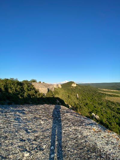 The image captures a scenic landscape with a prominent rock formation in the foreground and a lush green valley stretching into the distance under a clear blue sky. The rock in the foreground, appearing weathered and textured, casts a long shadow, suggesting the presence of a person or object not fully visible in the frame. The shadow's shape indicates it is a human figure, likely standing on the edge of the rock formation, looking out at the vista.
The mid-ground features a dramatic cliffside covered in dense green foliage and punctuated by large, light-colored rocks. These rocks rise above the trees, their surfaces illuminated by sunlight. Further in the distance, rolling hills covered in dark green trees recede into a hazy, verdant landscape. In the far distance, a lighter, cultivated field is visible, contrasting with the natural forest. The sky is a uniform, deep blue, free of clouds, indicative of clear weather.
The scene appears to be a natural park or mountainous region. Given the location provided, this view is likely from the Crimean Mountains near the settlement of Krasnomakskoye, Ukraine. The overall impression is one of natural beauty and a sense of vastness.