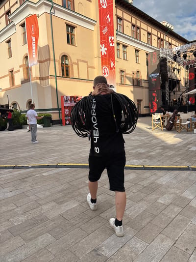 A man wearing black shorts and a black t-shirt with white lettering is carrying a large coil of black cables over his shoulders. He is standing on a paved ground surface with a yellow line marking a border. Behind him, a building with several windows and balconies is visible. A stage with lighting equipment and banners is set up in front of the building. Another person in a white t-shirt and grey pants is standing further away, looking at their phone. A red banner with white text is hanging on the building. The scene appears to be outdoors in a public space, possibly during a daytime event. The weather seems to be clear with sunlight casting shadows. Visible text on the man's shirt appears to be in reverse, possibly "HEADBORE 8". Text on a banner in the background reads "SING & PLAY". The overall atmosphere suggests a setup or preparation for an outdoor event.