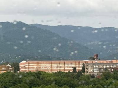 The image shows a city landscape with buildings and a mountainous backdrop. The foreground is dominated by lush green trees. Behind the trees, several buildings are visible. One prominent building is a large, long structure with a brick facade and horizontal white stripes. To its left, a smaller, lighter-colored building with visible windows is partially obscured. To the right of the brick building, a grey apartment building with multiple windows and balconies is present. In the distance, rolling hills and mountains covered in green foliage can be seen, with a cloudy sky overhead. The scene appears to be taken from an elevated viewpoint, possibly from a window. The image is slightly blurry and has several out-of-focus circular light spots, possibly water droplets on a window.