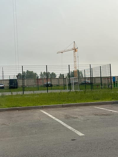 The image depicts a view of a construction site and a sports field, likely in the city of Novoaltaysk, Russia. In the background, a tall yellow construction crane stands against an overcast sky. In front of it, a chain-link fence encloses a concrete wall with graffiti. Within the fenced area, there is a small soccer field with a goal net. Several cars are parked behind the fence, some near a black van. The foreground shows a paved parking lot with white markings. Green grass and a curb line the edge of the parking lot. The scene appears to be captured during the daytime, under dull weather conditions.