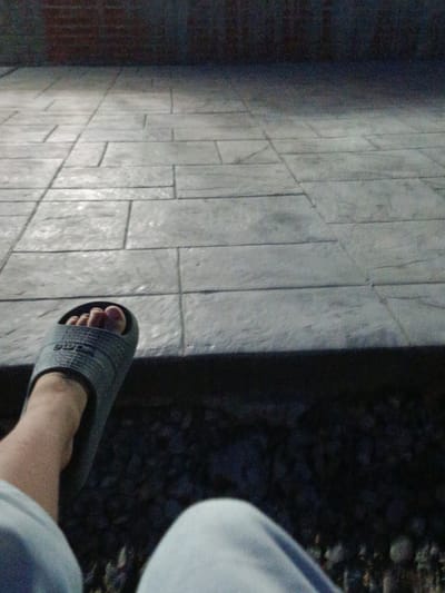 The image shows a close-up view of a person's foot wearing a gray slide sandal, resting on a tiled surface. The tiles appear to be rectangular and laid in a pattern. The background is dark and out of focus, with a hint of a brick wall at the top. The lighting suggests it might be dusk or night. The overall scene is of an outdoor area, possibly a patio or walkway. The location within Novotitarovskaya, Russia cannot be determined from the image.