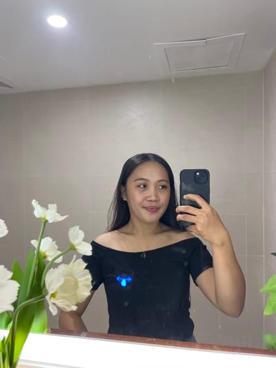 A young woman with long dark hair is taking a mirror selfie in what appears to be a bathroom. She is wearing an off-the-shoulder black top and a delicate gold necklace. In her right hand, she holds a black smartphone, which she is using to capture the image. To her left, a vase of white flowers is visible, their stems and leaves green. The background consists of tiled walls and a white ceiling with a circular light fixture and a ventilation grate. The mirror reflects the woman, the flowers, and the surrounding environment. The scene is clean and well-lit, suggesting a domestic or hospitality setting, likely within a residential area in Koronadal City, Philippines, given the presence of common household elements and the context provided.
