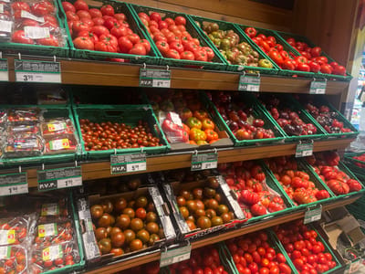 The image shows a supermarket produce aisle displaying a variety of tomatoes. The tomatoes are arranged in green plastic crates on wooden shelves. The varieties include large red tomatoes, some with ribbed surfaces, smaller red cherry tomatoes, and darker, multi-colored heirloom tomatoes. Price labels are visible above some of the crates, with prices indicated in Euros. One label mentions "P.V.P." which stands for "Precio de Venta al Público" (Retail Sale Price). The supermarket appears to be well-lit, and the overall impression is one of a clean and organized retail environment. There are no people visible in the immediate foreground, but the presence of multiple shelves filled with produce suggests a typical retail grocery store. The specific location in A Coruña, Spain, cannot be determined solely from the visual information in the image.