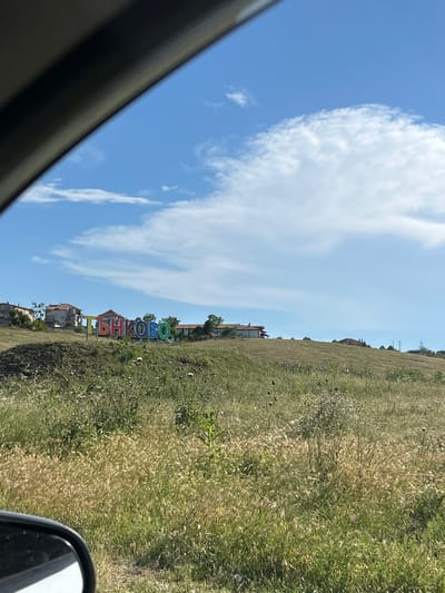 A brightly colored sign spelling "ТЪНКОВО" is situated on a grassy hill overlooking the town of Tankovo, Bulgaria. The letters are a vibrant mix of red, orange, yellow, green, and blue. Several houses with red-tiled roofs are visible in the background, nestled on the hillside. The sky is a clear blue with scattered white clouds. The foreground is filled with dry, tall grass and some green vegetation, with a dark car mirror visible in the lower left corner.
