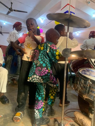 The image depicts a group of people gathered in what appears to be a musical setting in Nsugpe, Nigeria. Several individuals are focused on playing musical instruments, including a cymbal and drums. One child is wearing a shirt with colorful snowflake patterns. Other adults are present, some seemingly observing or assisting. The environment appears to be indoors, possibly a religious or community hall, judging by the decorations, including a flag and balloons. There is a focus on music performance.