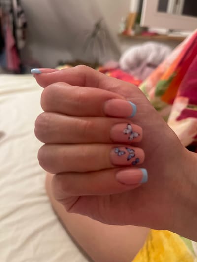 A hand with manicured nails is visible. The nails are painted in a pale pink color with a light blue French tip on the thumb, index, and pinky fingers. The middle and ring fingers are adorned with butterfly nail art. The hand is positioned against a blurred background of what appears to be bedding and a bedside table. The location of Saarbrücken, Germany is unknown.