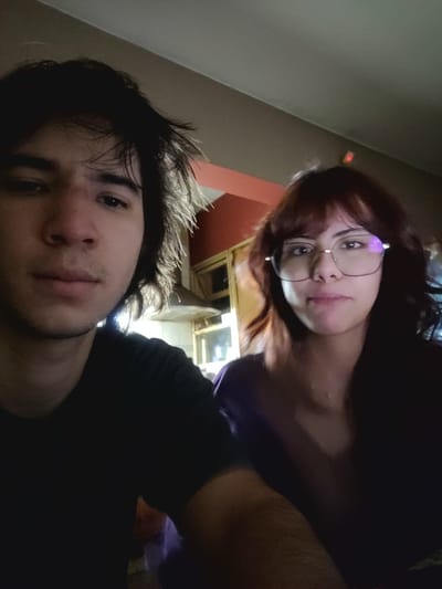 The image shows two people, one male and one female, likely in an indoor setting in San Fernando del Valle de Catamarca, Argentina. The male is in the foreground and has dark hair, light skin, and a slight beard. He is wearing a dark shirt. The female is in the background, slightly out of focus. She has reddish-brown hair, wears glasses, and appears to be wearing a purple top. The location is implied by the request and is not visually verifiable from the image alone. The image is tilted, with the top being on the left side and the bottom being on the right side. The lighting is dim. In the background, vague elements suggest an interior room with orange and beige walls.