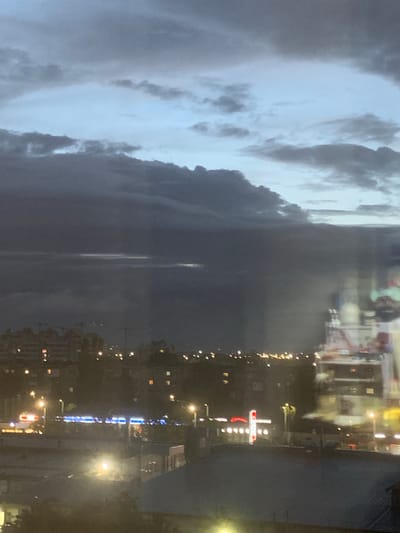This is a nighttime, aerial view of a city that is likely Kaliningrad, Russia, based on the included crop hints. The sky is filled with dark, heavy clouds with some lighter patches of sky visible. Below the clouds, the city lights are visible, creating a blurry glow. There are several buildings, some appearing to be apartment blocks with illuminated windows. Streetlights and signs are also visible, some with blue and red illumination. In the foreground, there are some darker building structures with roofs. There is a noticeable hazy or misty effect throughout the scene, possibly due to weather conditions or atmospheric effects. A tall, white structure with red detailing, possibly a building or a crane, is partially visible on the right side of the image. No people are clearly discernible in this image.