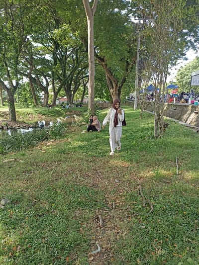 The image depicts a vibrant outdoor scene in a lush green park in Mataram, Indonesia, under bright daylight. In the foreground and midground, a woman walks towards the viewer, slightly to the right, smiling as she looks at a smartphone in her hands. She is dressed in a white long-sleeved shirt, light-colored pants, and a brown hijab, with a dark garment draped over her left arm. To her left, two other women are seated on the grassy bank of a calm stream or pond. One wears a black top and light-colored hijab, while the other wears a patterned top and a beige hijab, both appearing relaxed.
The background is dominated by numerous large, mature trees with dense green foliage. Across the stream, several groups of people are seated on the grass under the trees, suggesting a social gathering or general relaxation. On the far right side of the background, an outdoor market or community event is visible, featuring blue tents or canopies, people, and stalls with various goods, alongside colorful buckets on a low wall. A utility pole is also present in this area. The ground is covered in green grass with scattered dry leaves. On one of the distant blue tents, a partial, indistinct letter "И" or a similar stylized character is faintly visible. The scene suggests a pleasant day for leisure and community activity in a public park.