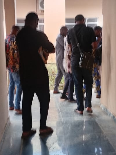 The scene depicts approximately eight adult males gathered within a brightly lit, tiled corridor or covered walkway in Minna, Nigeria. In the immediate foreground, two men are viewed from behind, exhibiting slight motion blur. The figure on the left wears a dark long-sleeved shirt, dark trousers, and brown shoes; his arms appear folded or he holds an obscured object. The figure on the right wears a dark short-sleeved polo shirt, dark trousers, light-colored shoes or sandals, and carries a dark messenger bag over his right shoulder. Behind these individuals, several other men are visible, dressed in a mix of traditional patterned attire, a light grey traditional garment, and Western casual wear. The environment features smooth, light beige walls, a highly reflective grey tiled floor, and an exterior air conditioning unit visible in the upper background through an opening. Green foliage is discernable beyond this opening. No specific actions or direct interactions are clearly identifiable due to the image's overall blur.