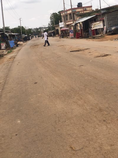 A street scene in Hanwa, Nigeria. The paved road, heavily pitted with potholes, dominates the frame. A single person is walking on the right side of the road, away from the camera. The person is wearing a white top and dark pants. The roadside contains a mix of one and two-story buildings. There are commercial buildings/shops visible. Powerlines and utility poles are also visible. The sky is overcast.